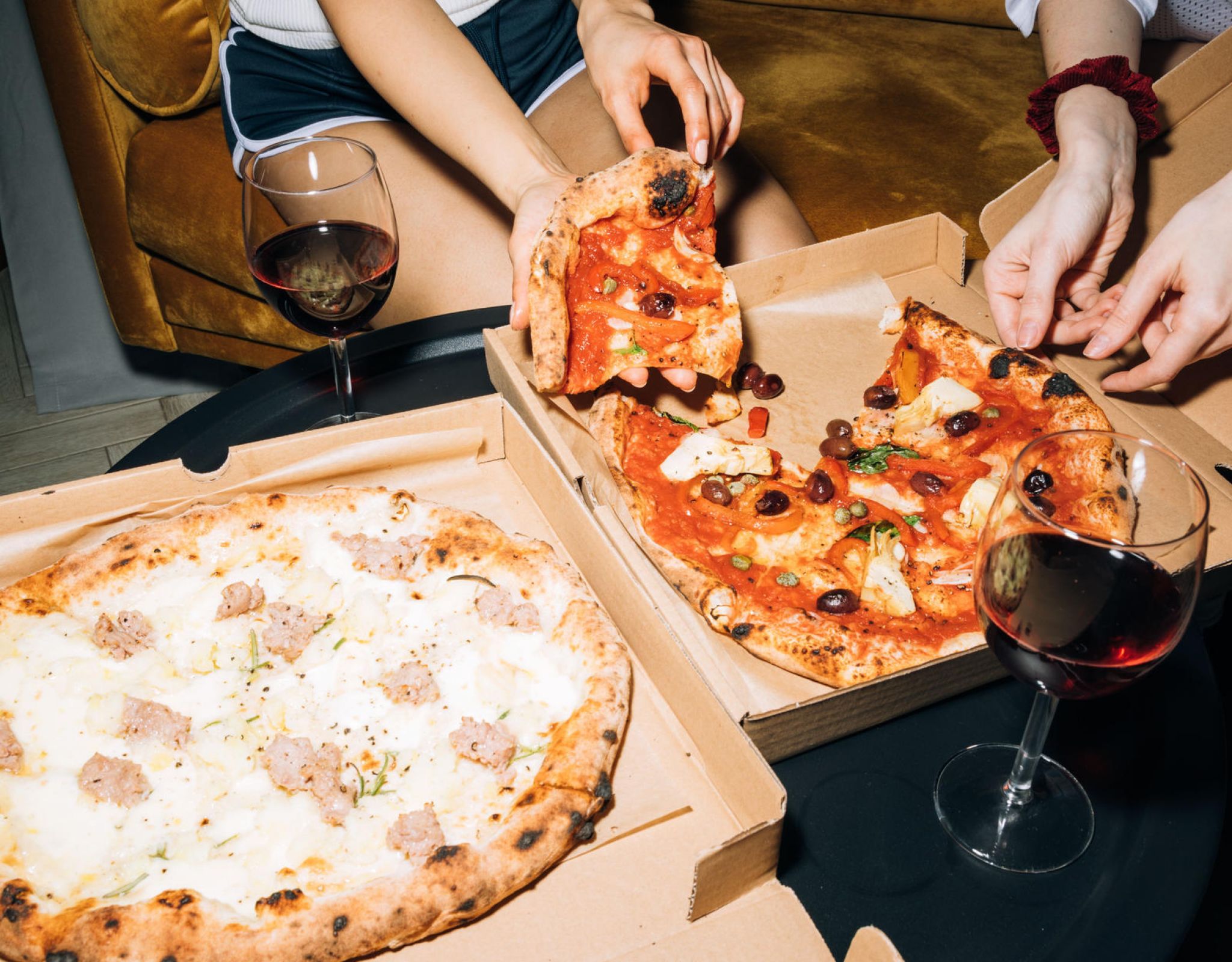 Two people share pizza and red wine at a table, with two open pizza boxes and glasses of wine in front of them.