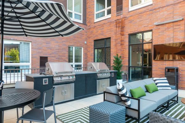 A modern outdoor grill area with shaded picnic tables and stainless steel barbecue stations surrounded by landscaped greenery at Chronicle on 8th.