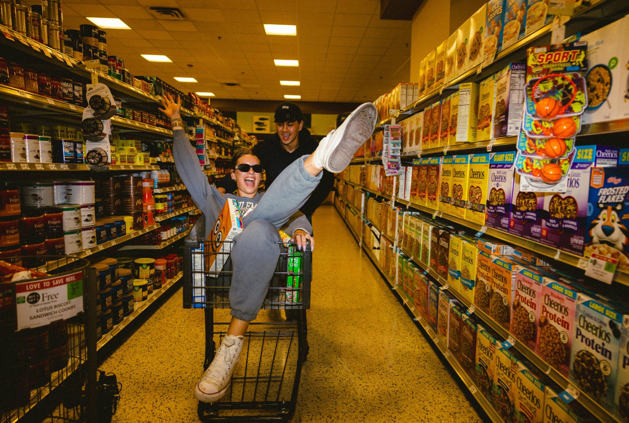 Two people in a grocery store aisle, one pushing the other, who is laughing and sitting in a shopping cart with arms and legs raised.