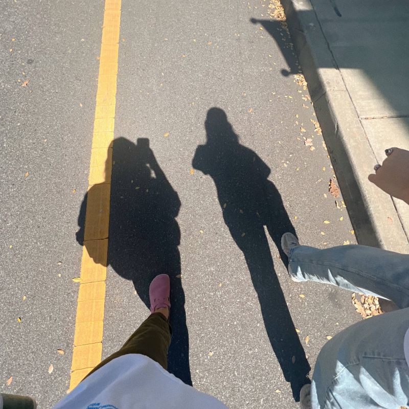 Three people’s shadows cast on a sunlit street with a yellow line, feet and legs partially visible.