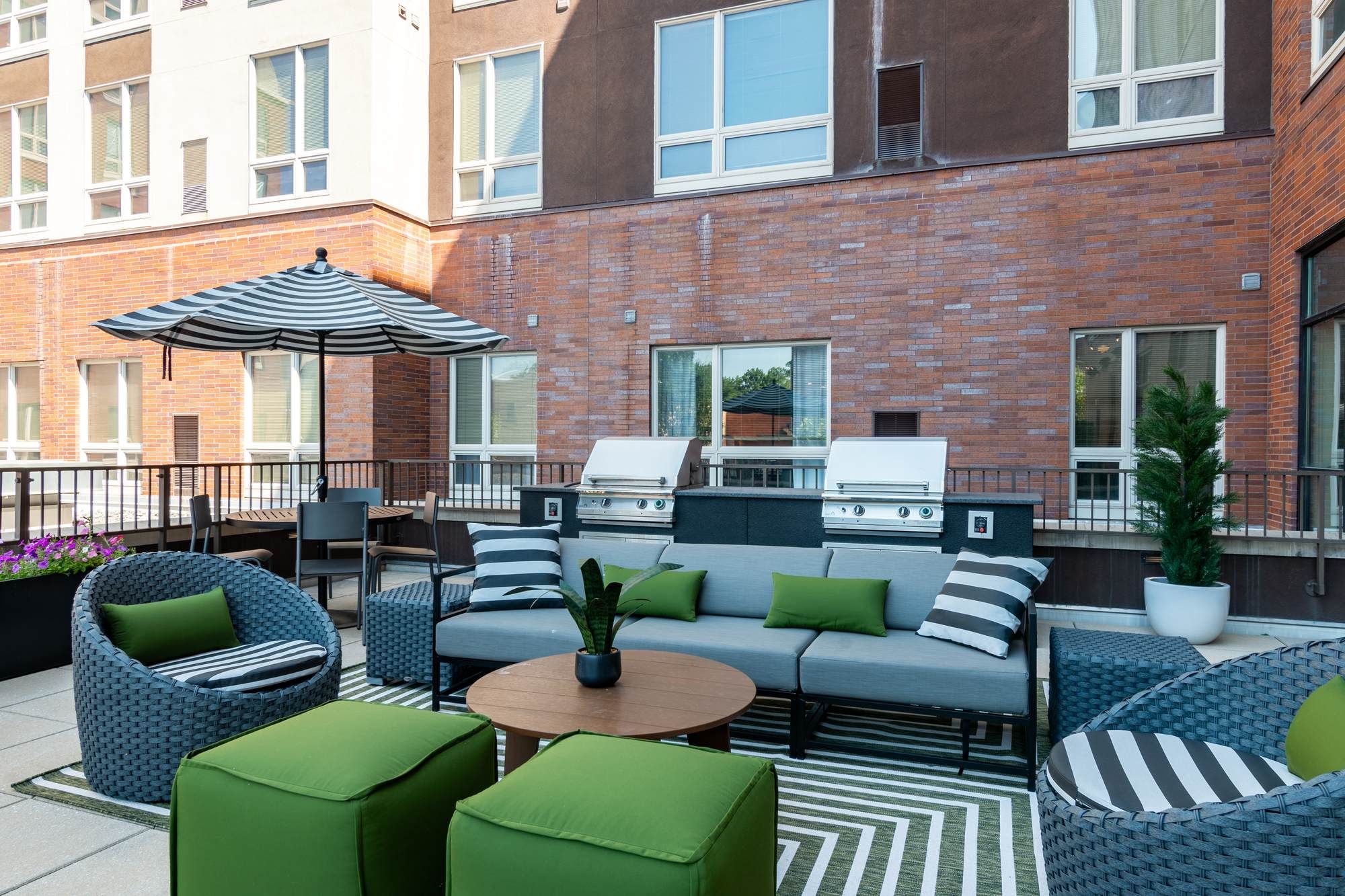 Outdoor lounge area at Chronicle on 8th featuring modern patio furniture, green accent cushions, umbrellas, and built-in grills against a brick exterior backdrop.