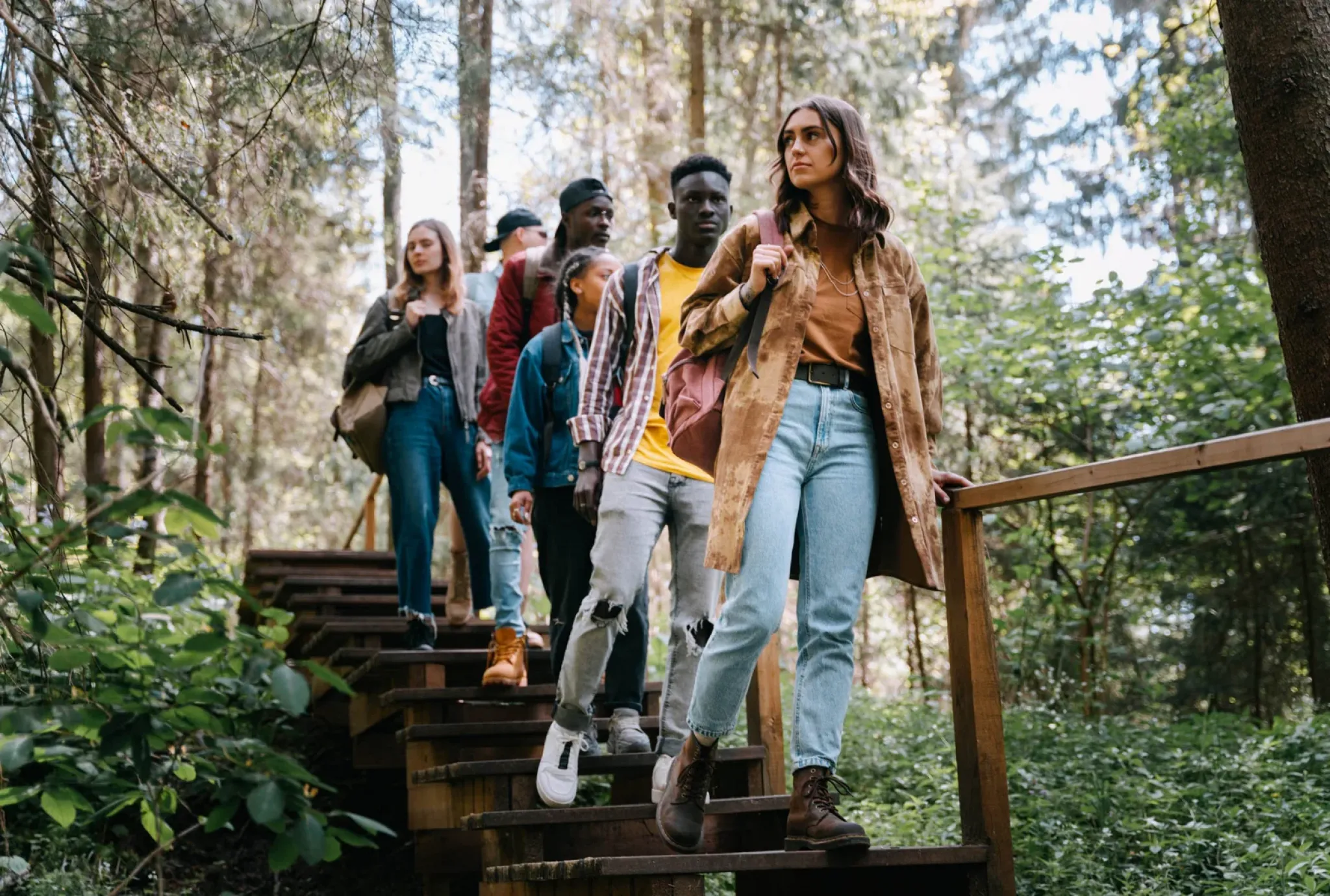 A group of young adults hikes down wooden steps in a lush green forest, surrounded by trees and sunlight.