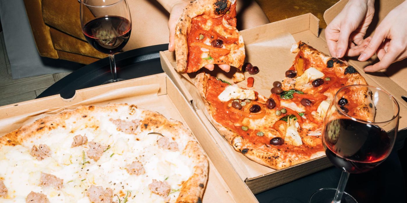 Two people share pizza and red wine at a table, with two open pizza boxes and glasses of wine in front of them.