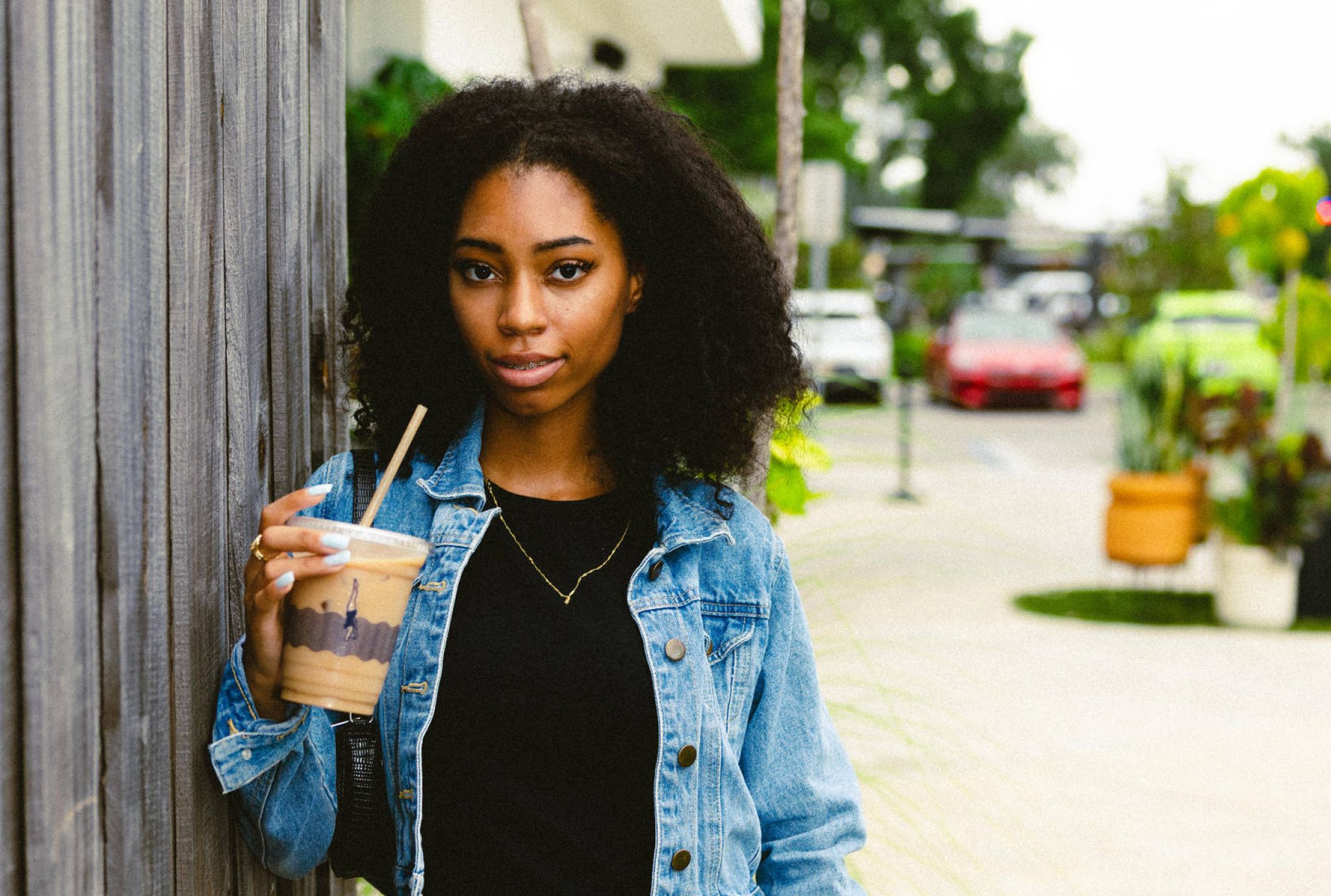 Woman in a denim jacket and orange skirt holds iced coffee, standing by a wooden fence on a city sidewalk.
