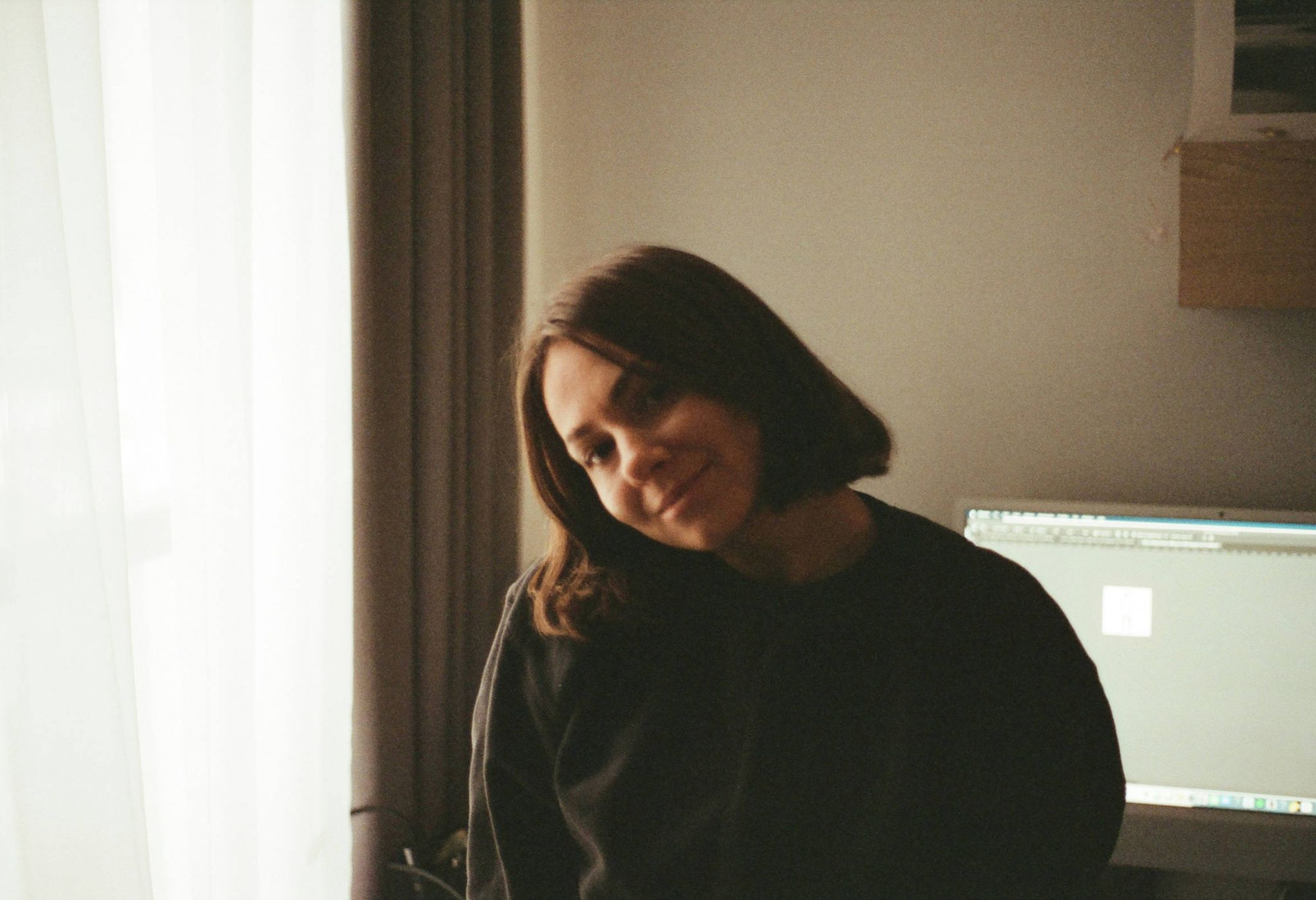 A person with short brown hair smiles softly while sitting in a softly lit room near a computer monitor.
