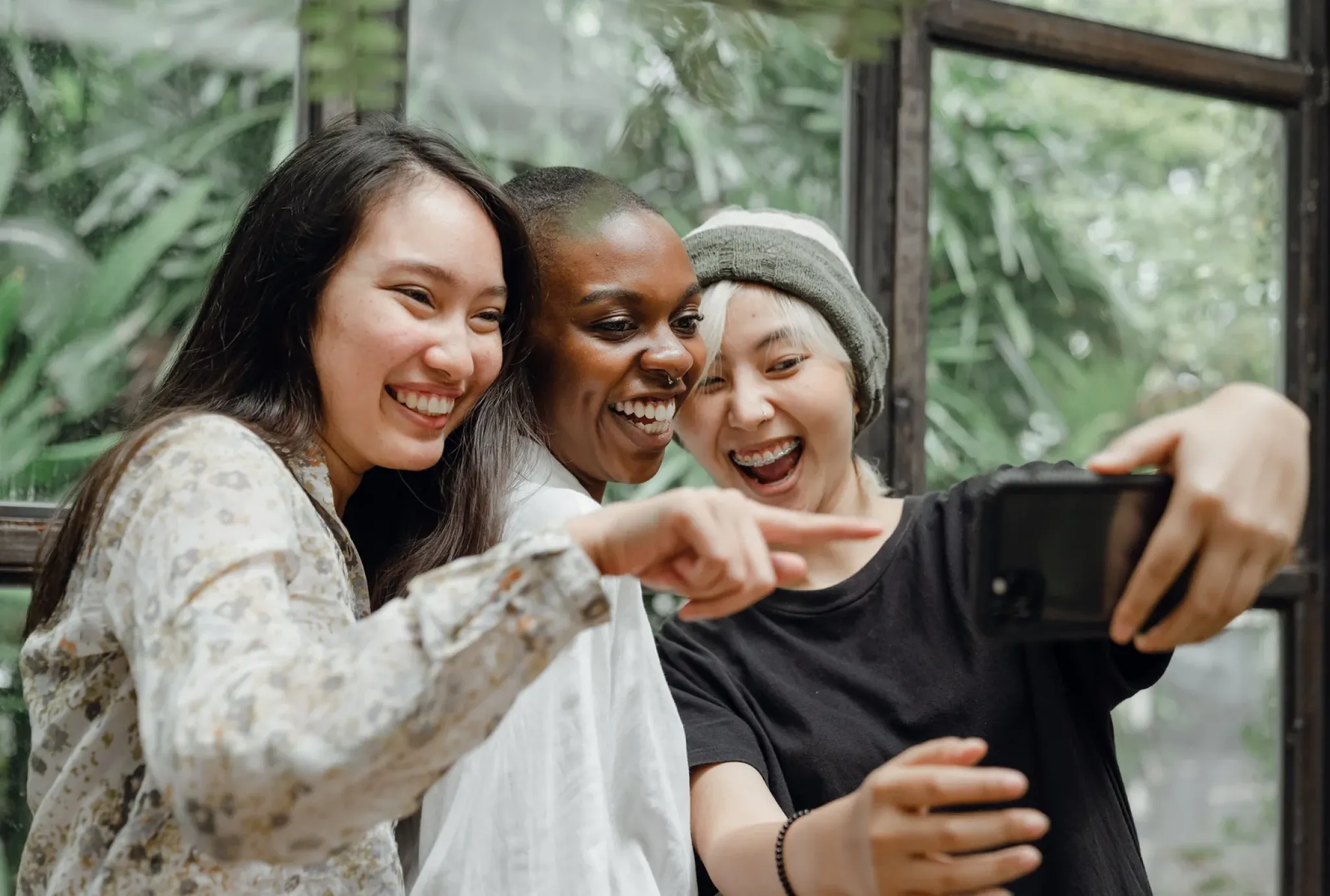 Three friends smiling and taking a selfie together indoors, with greenery visible through the windows behind them.