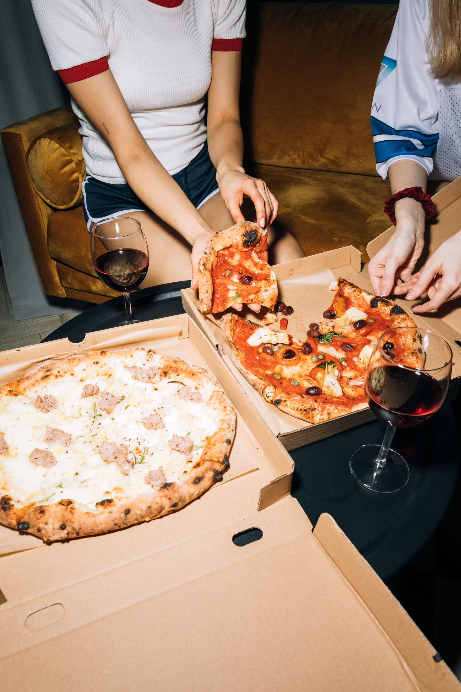 Two people share pizza and red wine at a table, with two open pizza boxes and glasses of wine in front of them.