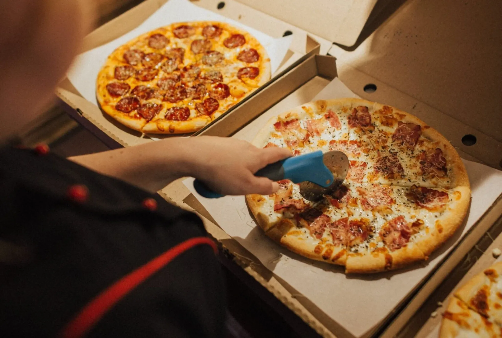 A person uses a pizza cutter to slice a pizza with ham and herbs, with another pizza in a box nearby.