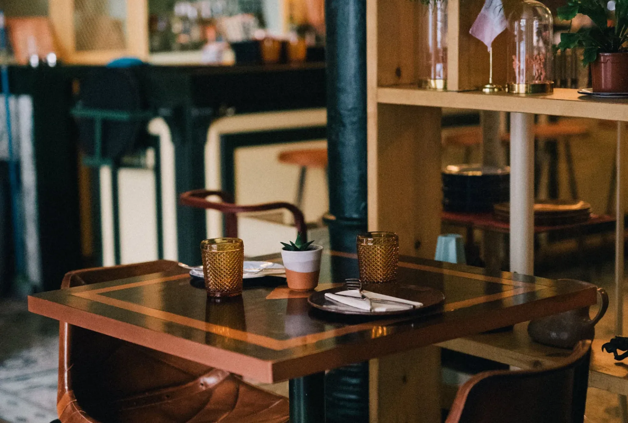 A small restaurant table with two glasses, plates, cutlery, and a potted plant, set in a cozy, dimly lit interior.