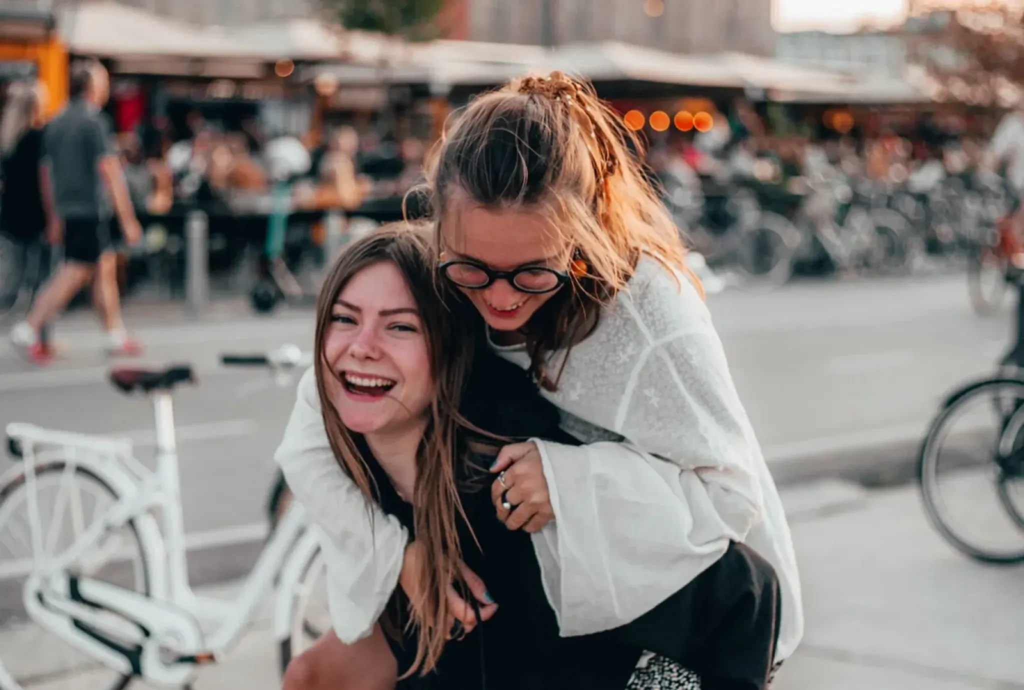 Two young women laughing, one giving the other a piggyback ride on a city street near outdoor cafes and parked bicycles.