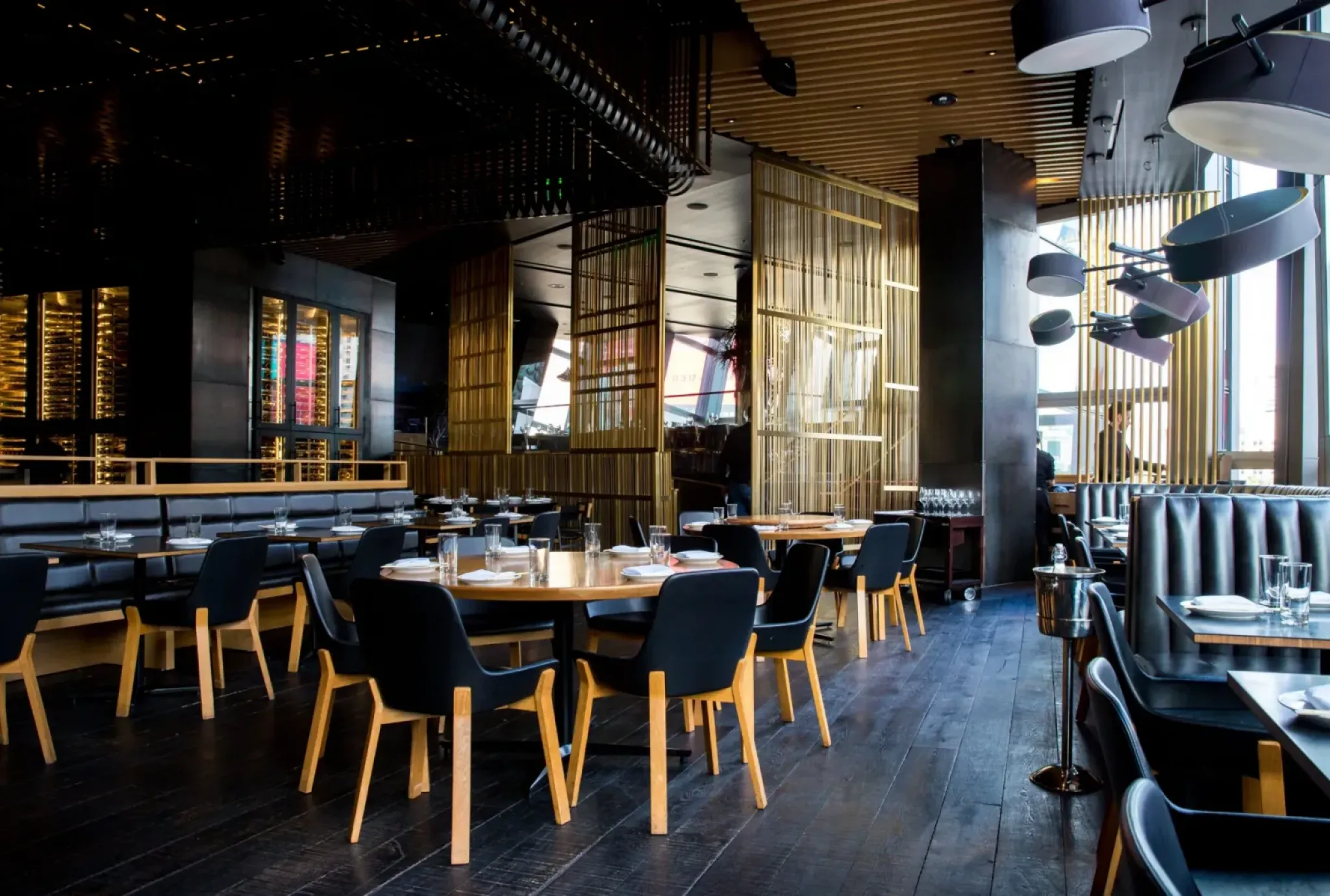 Modern restaurant interior with wooden tables, black chairs, set place settings, and large windows letting in natural light.