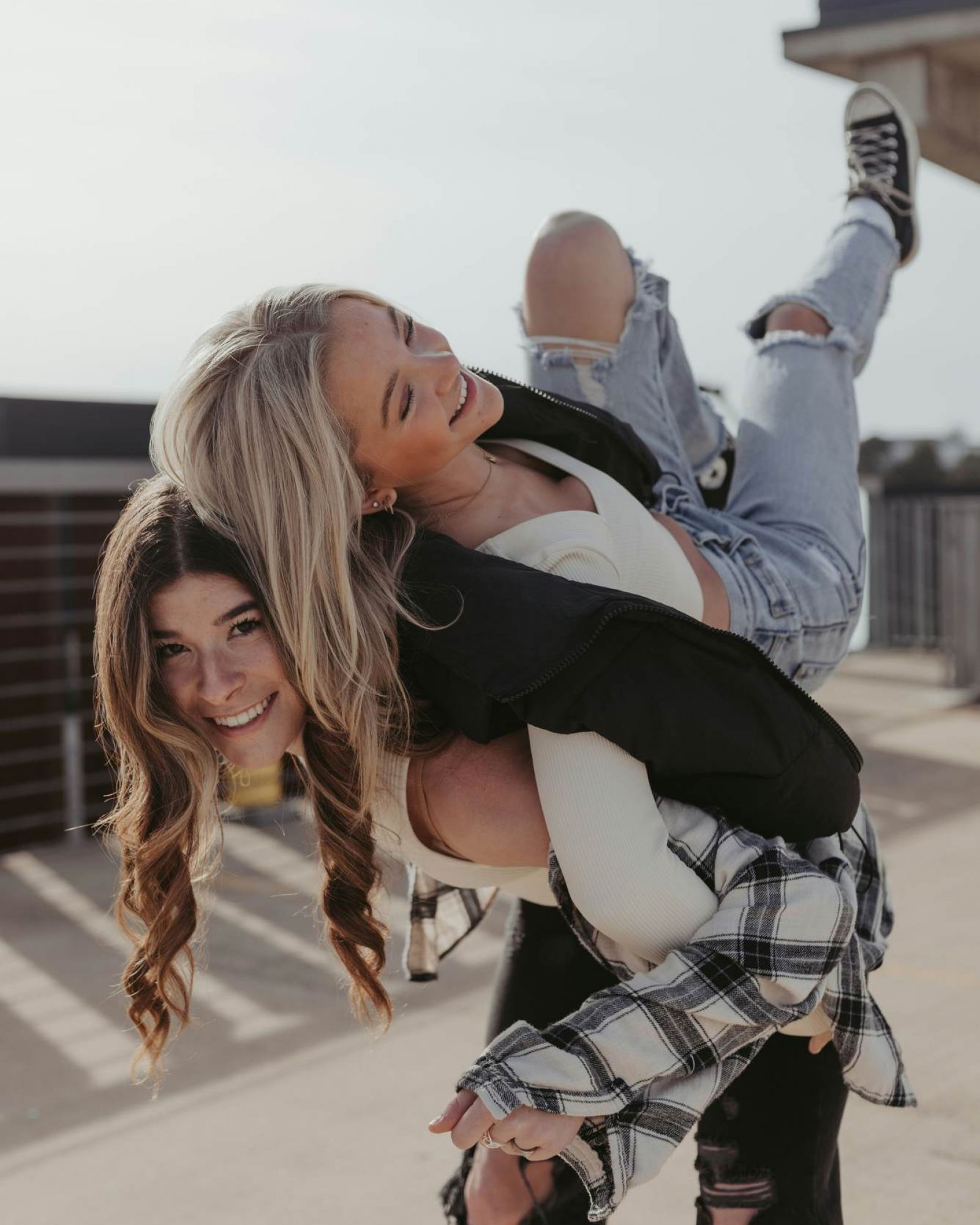 Two young women laugh as one gives the other a playful piggyback ride on a rooftop, both wearing casual ripped jeans.