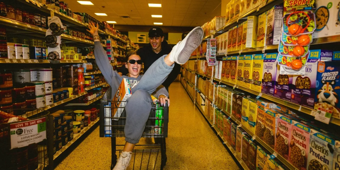 Two people laugh as one sits in a grocery cart with arms raised, pushed by the other down a brightly lit cereal aisle.