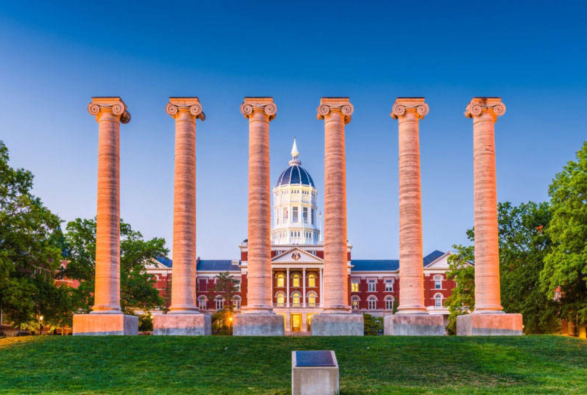 Six stone columns stand on grass in front of a historic red-brick building with a dome, under a clear blue sky.