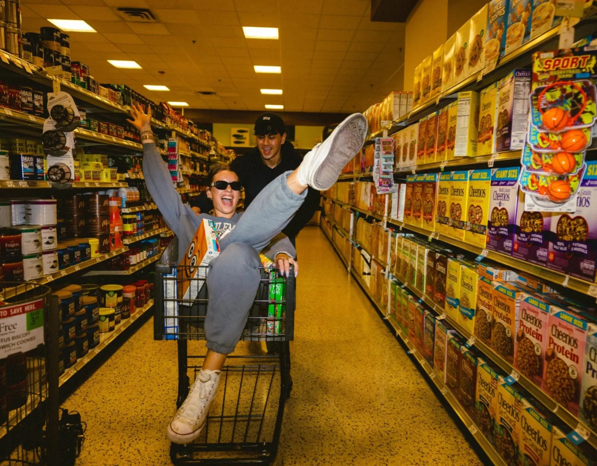 Two people laugh as one sits in a grocery cart with arms raised, pushed by the other down a brightly lit cereal aisle.